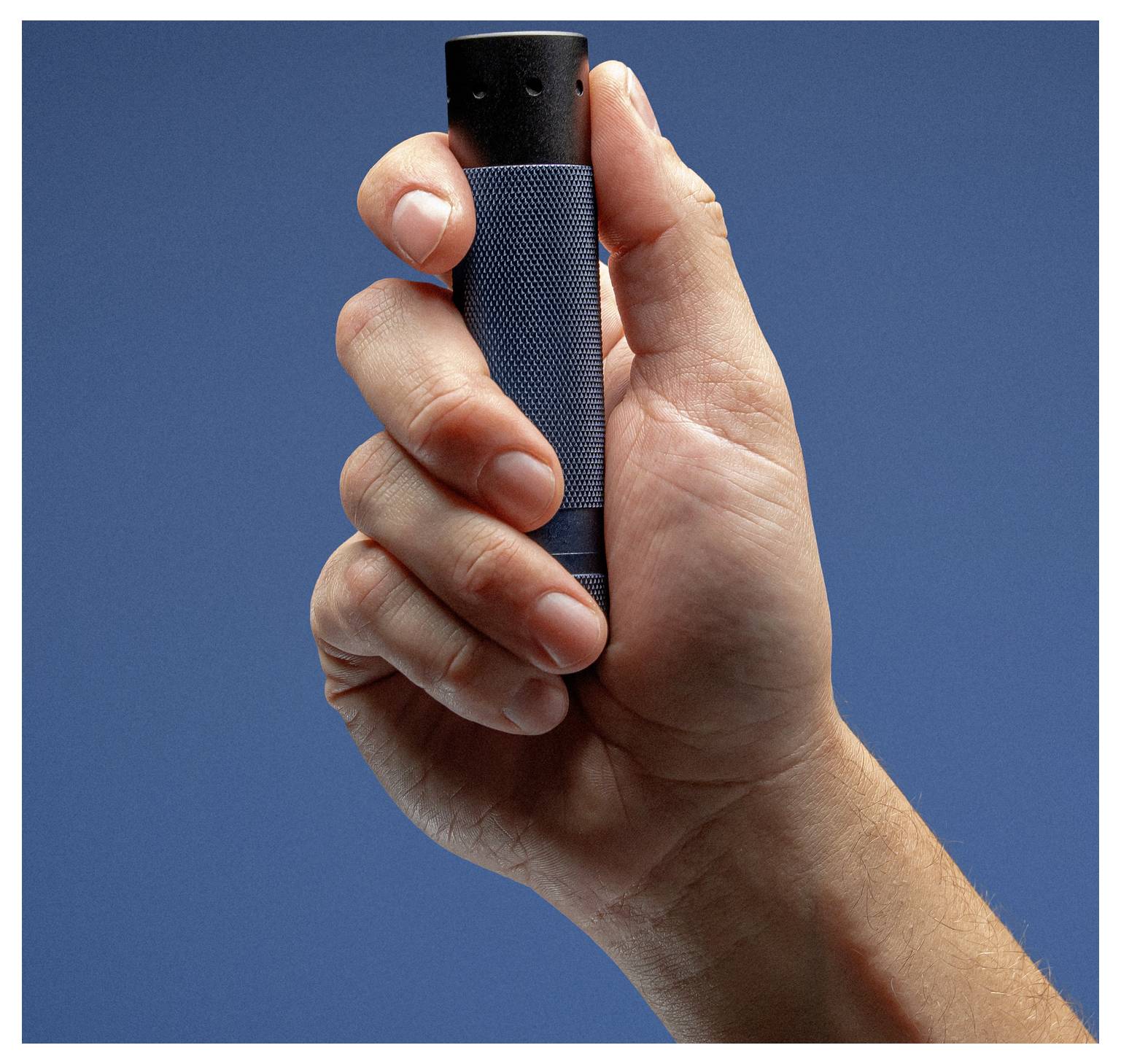 Eine Person hält ein kleines zylindrisches Gerät mit strukturierter Griffoberfläche gegen einen einfarbig blauen Hintergrund, was auf praktische Anwendung oder Techniknutzung hindeutet.