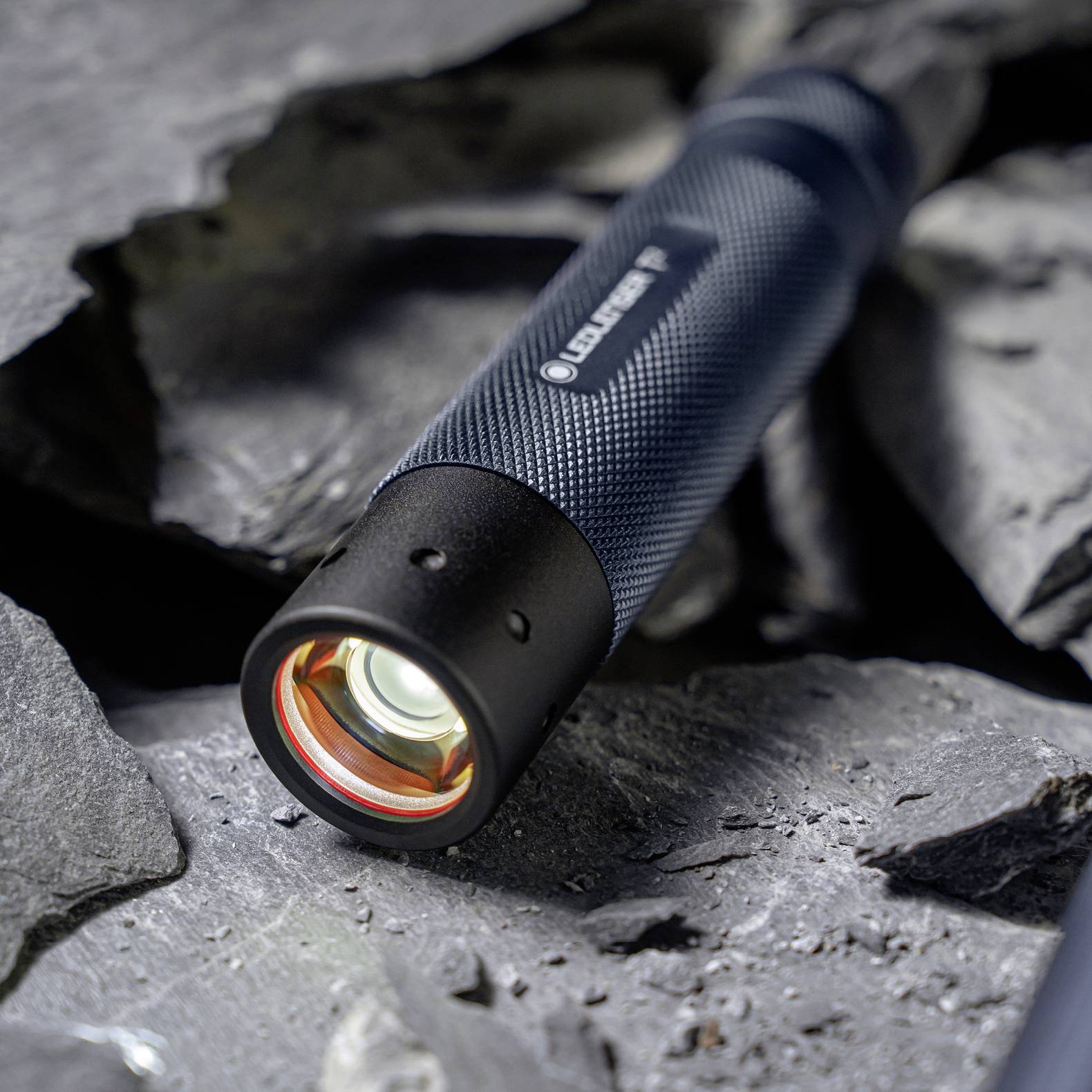 Eine Taschenlampe mit leuchtendem Objektiv ruht auf dunklen, strukturierten Schiefersteinen. Die Taschenlampe hat einen schwarzen, strukturierten Griffgriff.