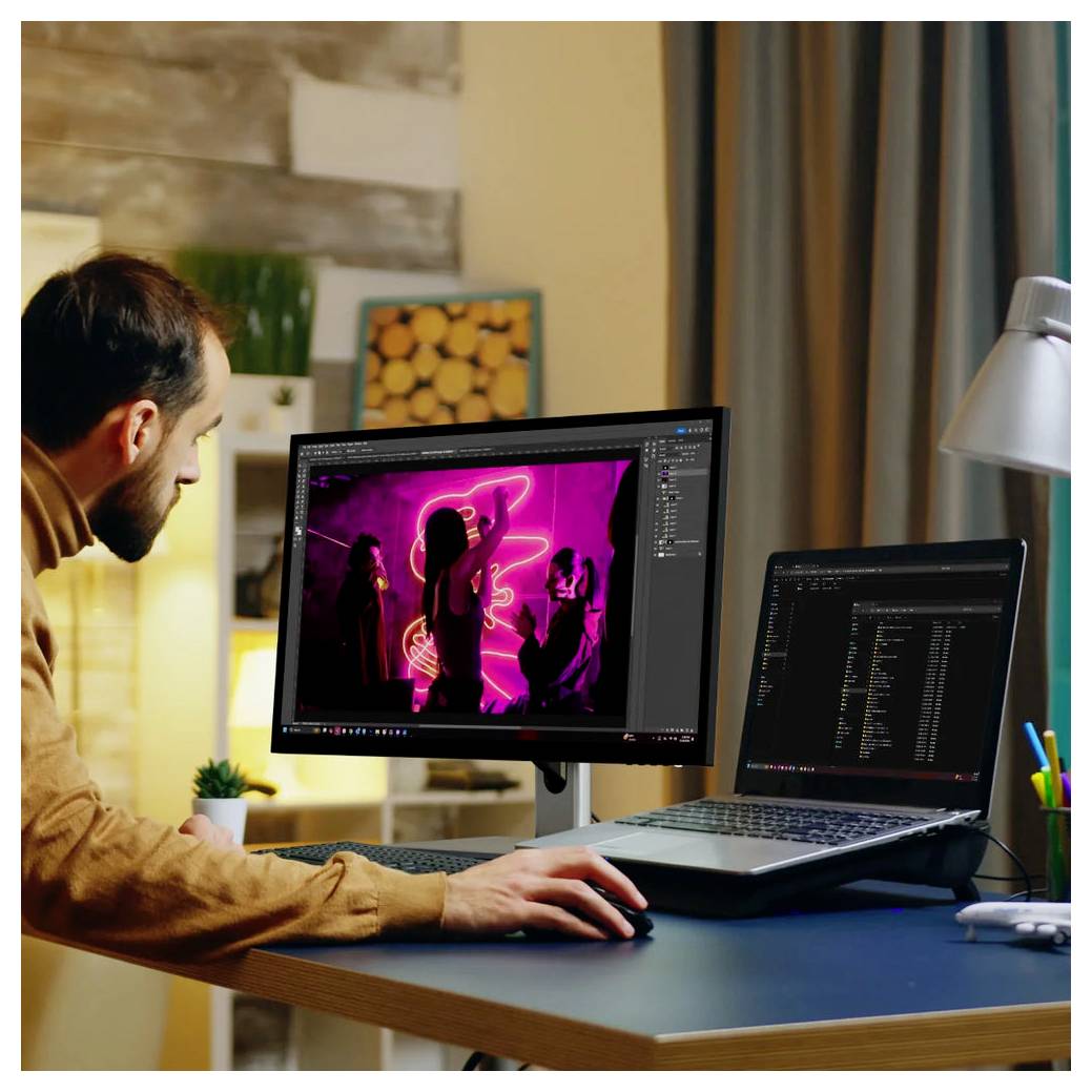 Ein Mann bearbeitet ein neonbeleuchetes Foto auf einem Desktop-Monitor, während er einen Laptop an einem Schreibtisch in einem Homeoffice nutzt.