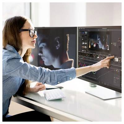 Frau bearbeitet Video auf zwei Monitoren, zeigt auf eine Timeline und Vorschau eines Männerporträts in Farbgrading-Software.