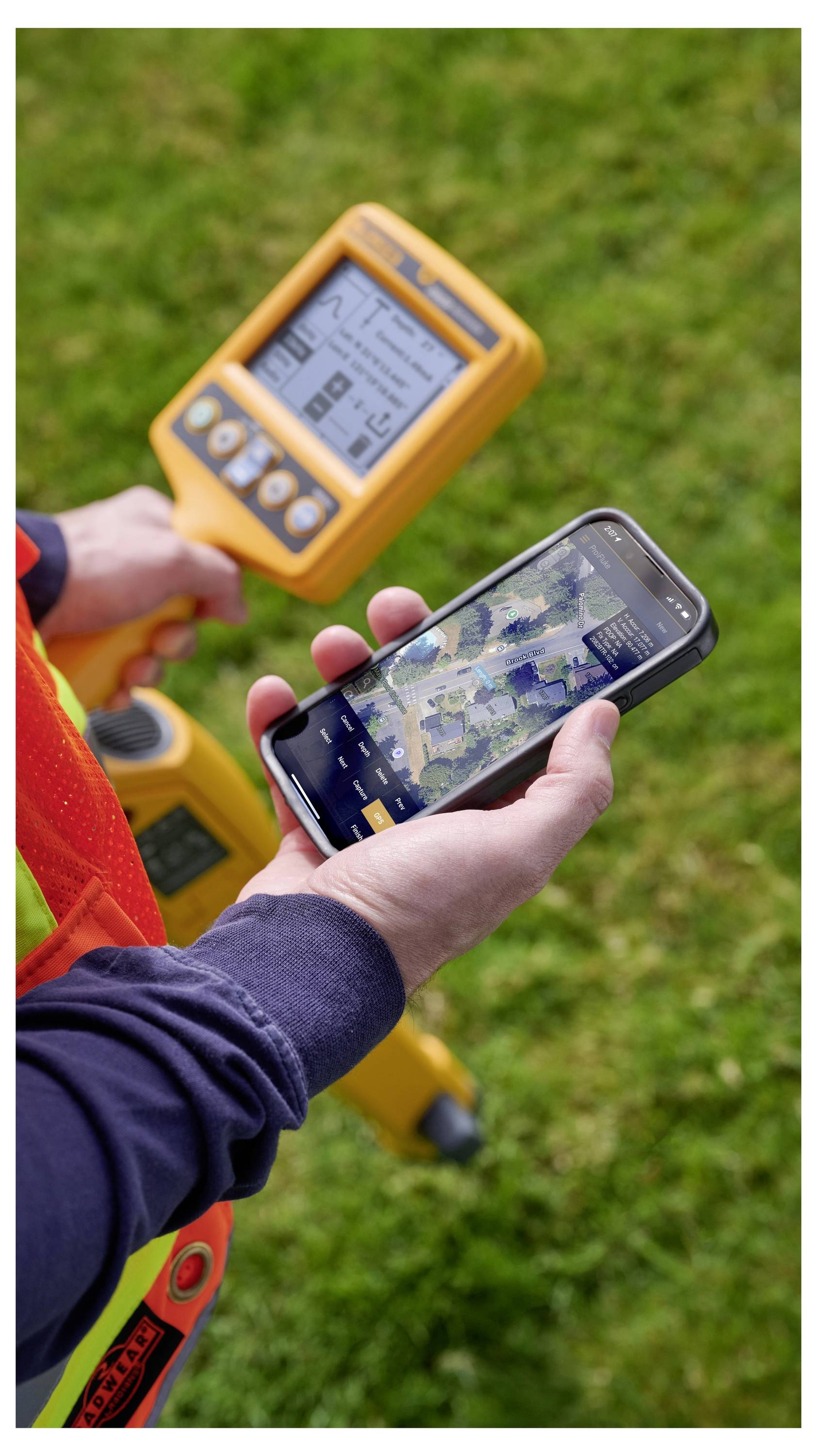 Person an einer Außenbaustelle nutzt ein Smartphone neben gelben Vermessungsinstrumenten auf einer Grünfläche.