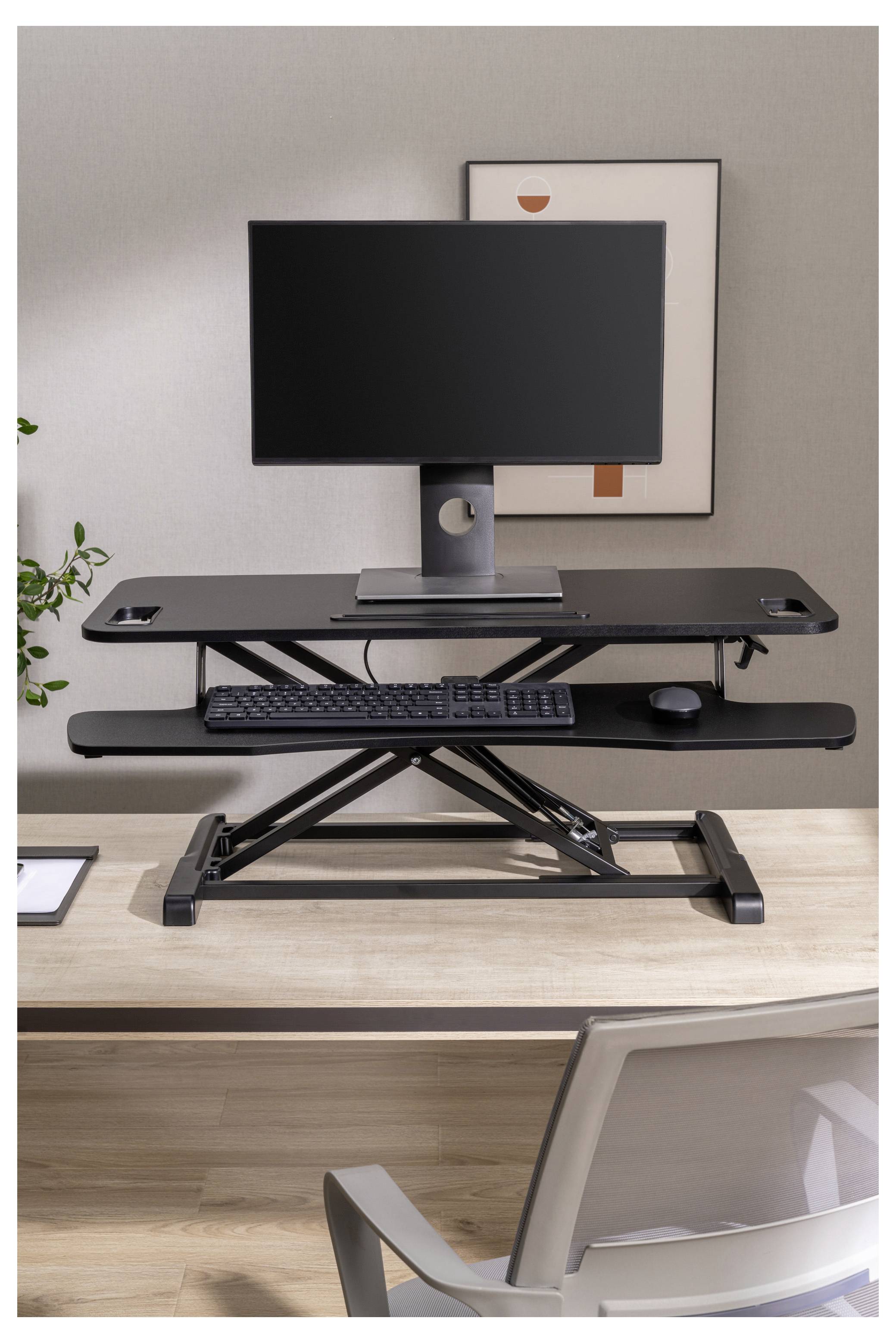 Ein verstellbarer Stehschreibtisch mit einem Monitor, einer Tastatur und einer Maus steht auf einem Holzschreibtisch in einem modernen Büro, mit einem Stuhl im Vordergrund.
