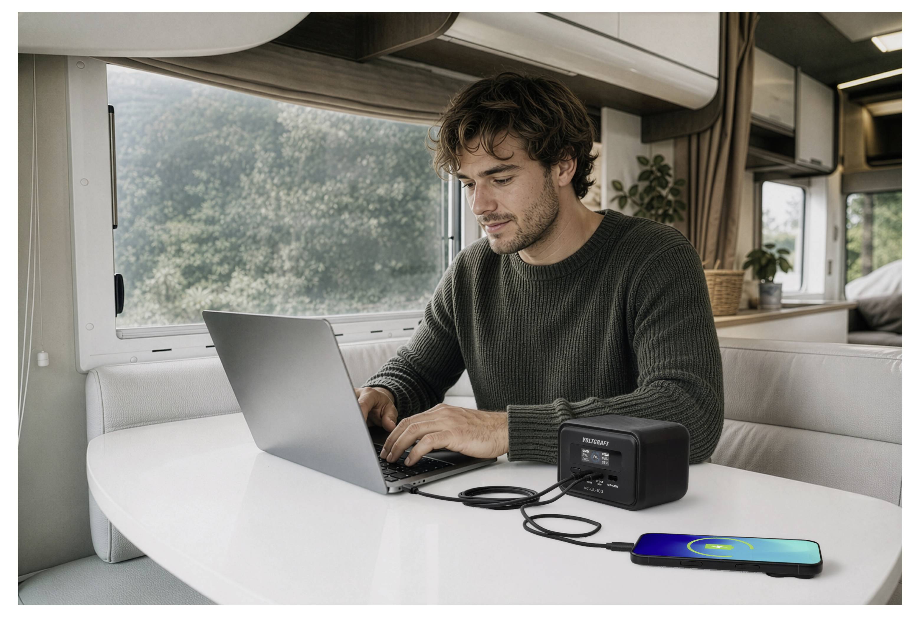 Mann, der einen Laptop an einem Tisch in einem Wohnmobil benutzt, mit einer tragbaren Stromversorgung und einem Smartphone in der Nähe, das gerade aufgeladen wird.