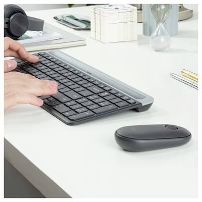 Hände tippen auf einer schwarzen kabellosen Tastatur an einem weißen Schreibtisch mit Maus, Kopfhörern, Notizbuch, Stiften und Sanduhr.