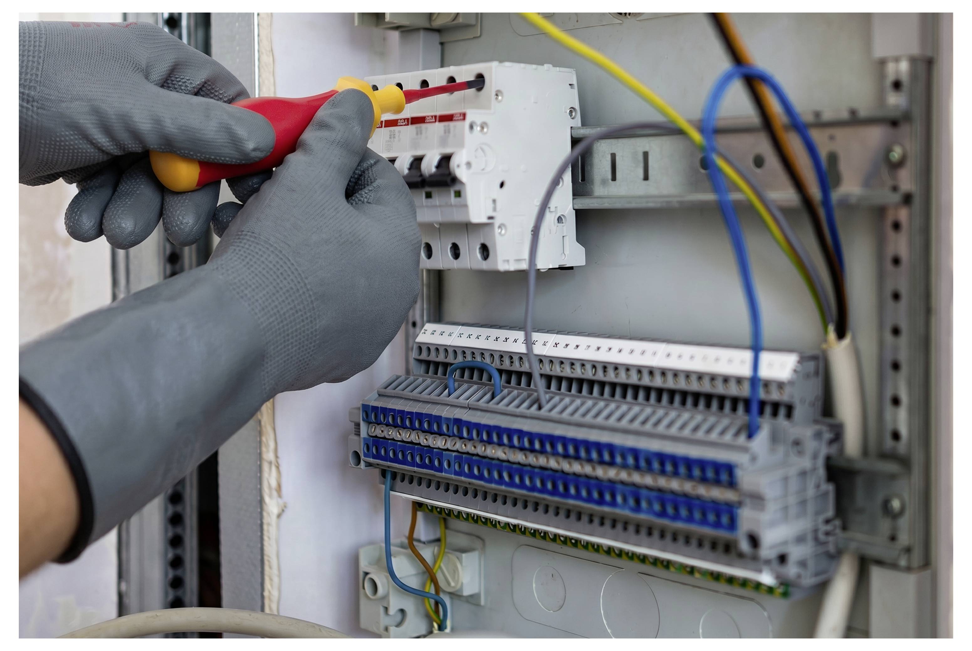 Handschuhgetragener Elektrotechniker, der einen Schutzschalter in einem Stromverteilerschrank mit angeschlossenen Drähten und Anschlussblöcken festzieht.