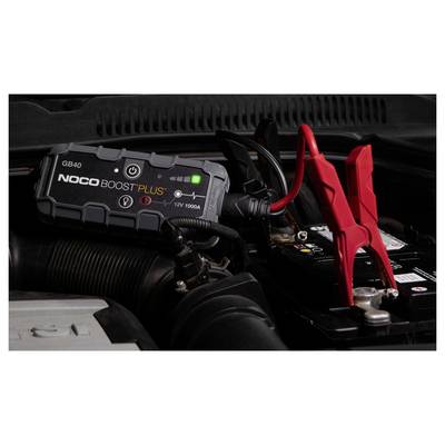 Ein NOCO Boost Plus Starthilfegerät ist mit roten und schwarzen Klemmen an einer Autobatterie befestigt und zeigt den Motorraum des Fahrzeugs.