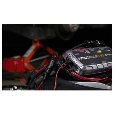 Nahaufnahme eines NOCO Boost Pro Starthilfegeräts mit Kabeln und einer behandschuhten Hand neben einer Autobatterie in einem Motorraum.