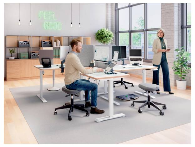 Modernes Großraumbüro mit einem Mann, der an einem Schreibtisch arbeitet, und einer Frau, die in der Nähe steht; ein leuchtendes "FEEL GOOD"-Schild an der Wand.