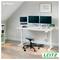 Homeoffice mit verstellbarem Schreibtisch, Dual-Monitoren, Laptop, Hocker, Tastatur und Pflanze unter gerahmter Wandkunst; "Zu Hause" und LEITZ-Logo sichtbar.