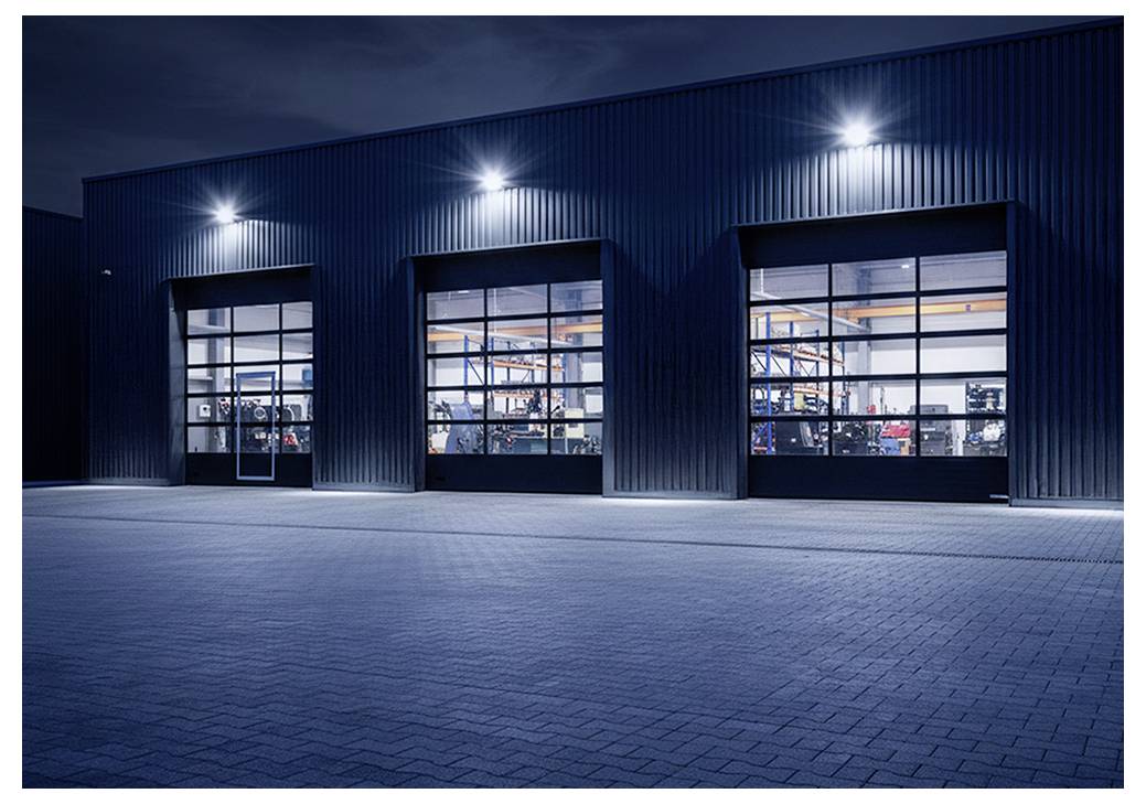 Industrielles Lagerhausgelände bei Nacht mit drei beleuchteten gläsernen Garagentoren und einem gepflasterten Vorplatz.