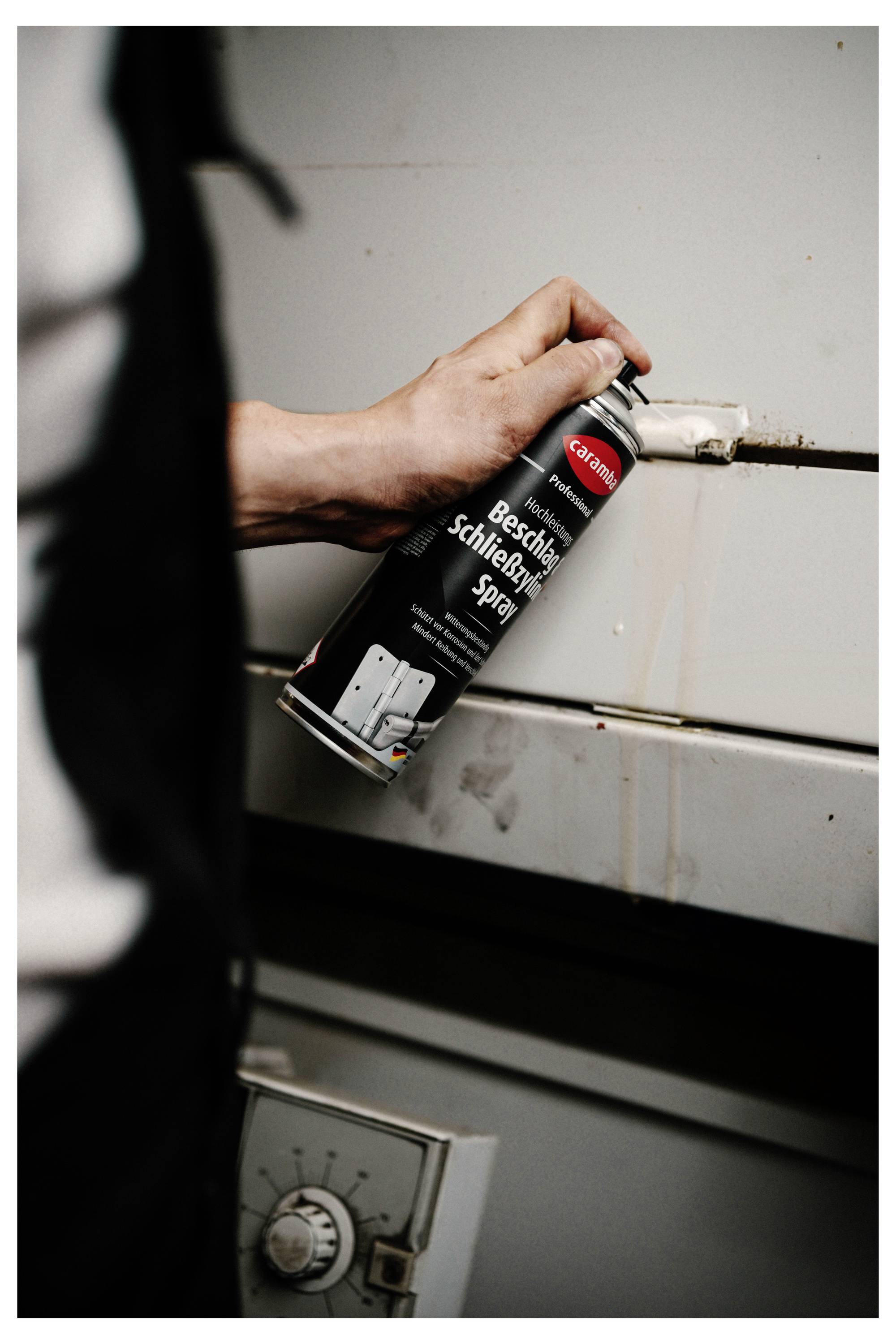 Hand sprüht Schmiermittel aus einer CRC 'Beschlag Schmierspray'-Dose auf ein Metallscharnier oder Schloss an einem grauen Schrank oder einer grauen Tür.