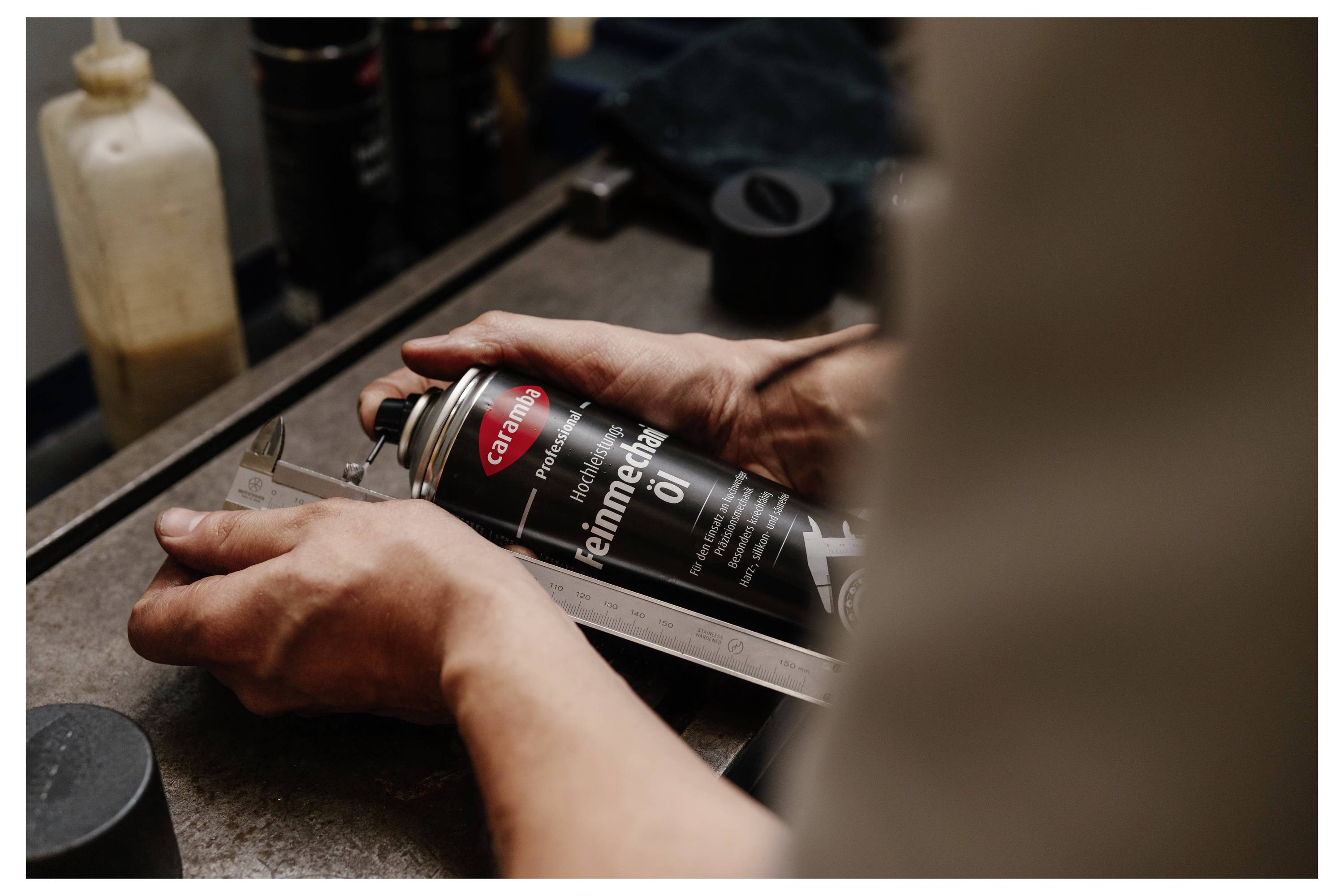 Eine Dose Caramba Professional Hochleistungs-Präzisionsöl steht auf einem Werkstatttisch neben Metallteilen, mit unscharfem Industriehintergrund.