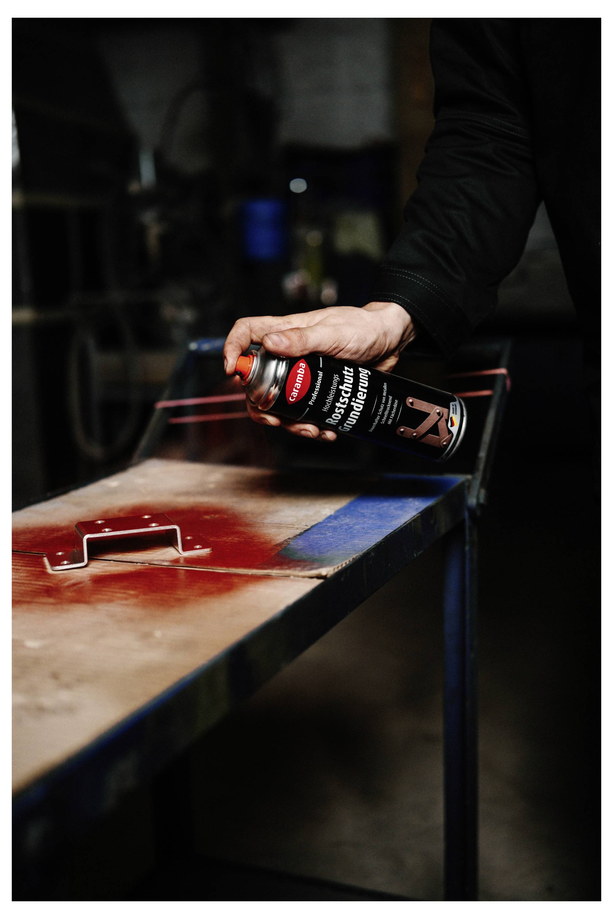 Eine Person sprüht rote Farbe aus einer Sprühdose auf eine Metalloberfläche in einer Werkstatt.