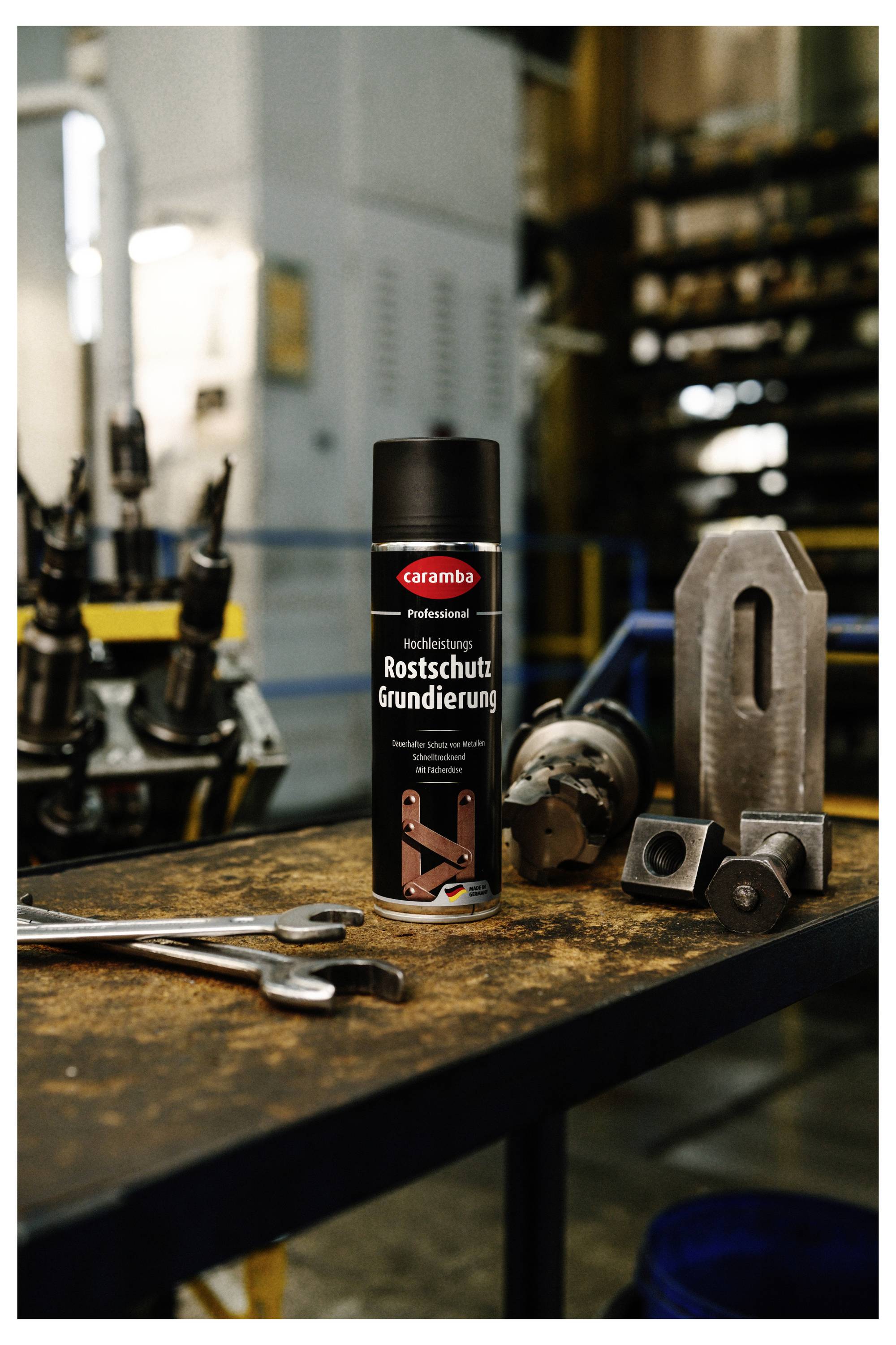 Sprühdose mit der Aufschrift "Caramba Rostschutz Grundierung" auf einem Werkstatttisch mit Metallwerkzeugen und Maschinenteilen, was auf eine Rostschutz-Grundierung hinweist.