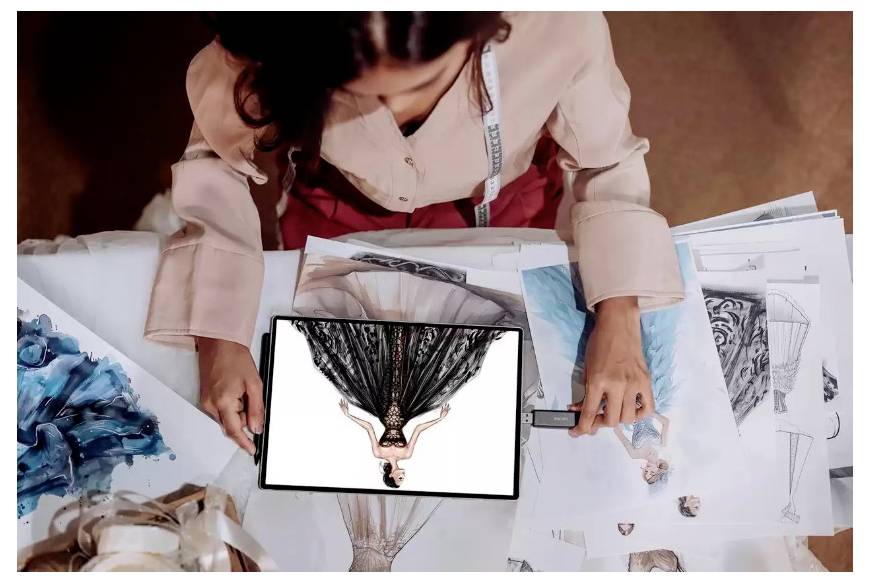Modedesigner ordnet Skizzen und Stoffmuster um ein Tablet herum an, das ein Kleidungsdesign zeigt.