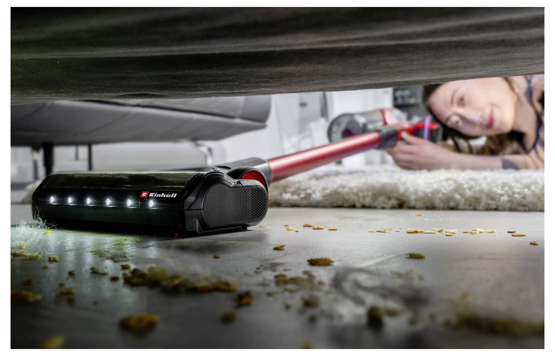 Eine Frau reinigt unter einem Bett mit einem roten kabellosen Stabstaubsauger und erreicht Schmutz und Krümel in einem niedrigen, schwer zugänglichen Raum.