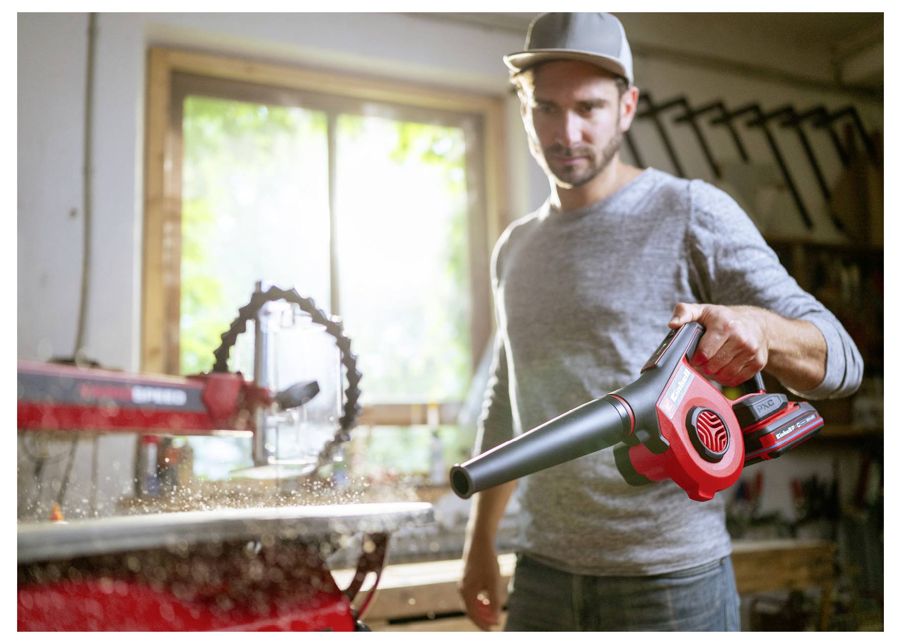 Ein Mann benutzt einen roten kabellosen Laubblaser in einer Werkstatt und bläst Staub und Ablagerungen von einer Werkbank neben einem Fenster weg.