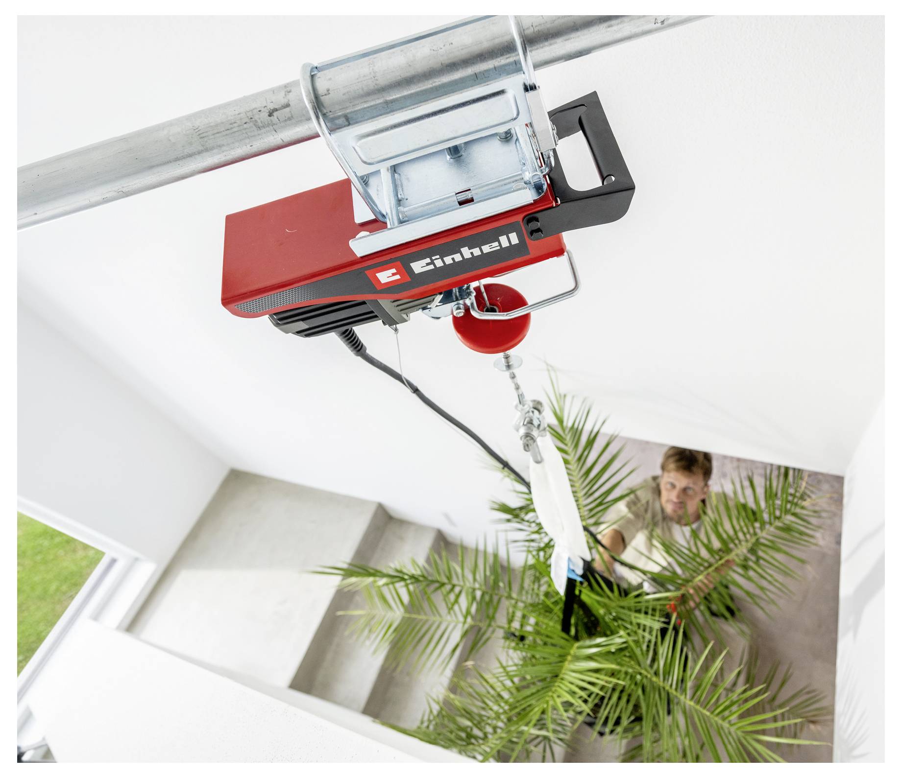 Ein roter elektrischer Deckenhebezug hebt eine Pflanze durch ein Treppenhaus, während eine Person sie von unten lenkt.