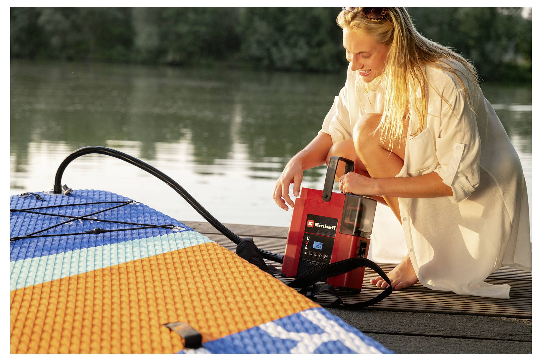 Eine Frau kniet auf einem Steg neben einem Paddelbrett und benutzt eine tragbare Pumpe, um es neben einem See aufzupumpen.