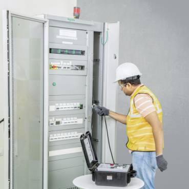 Arbeiter in Schutzhelm und Warnweste inspiziert eine offene Elektroschalttafel mit tragbarem Prüfgerät und Ausrüstungskoffer.