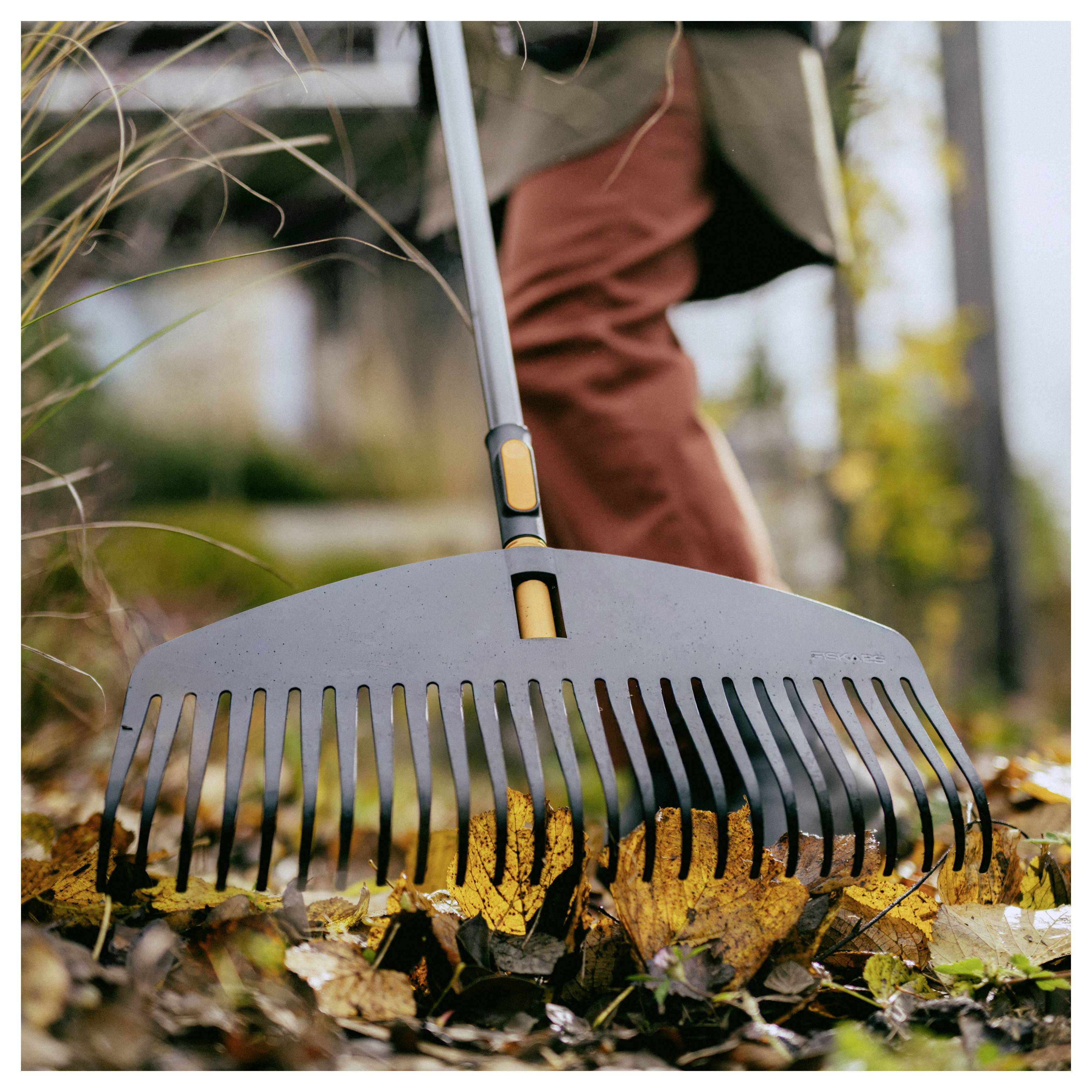 Person, die mit einem breiten Laubrechen gefallene Herbstblätter in einem Garten zusammenrechen.
