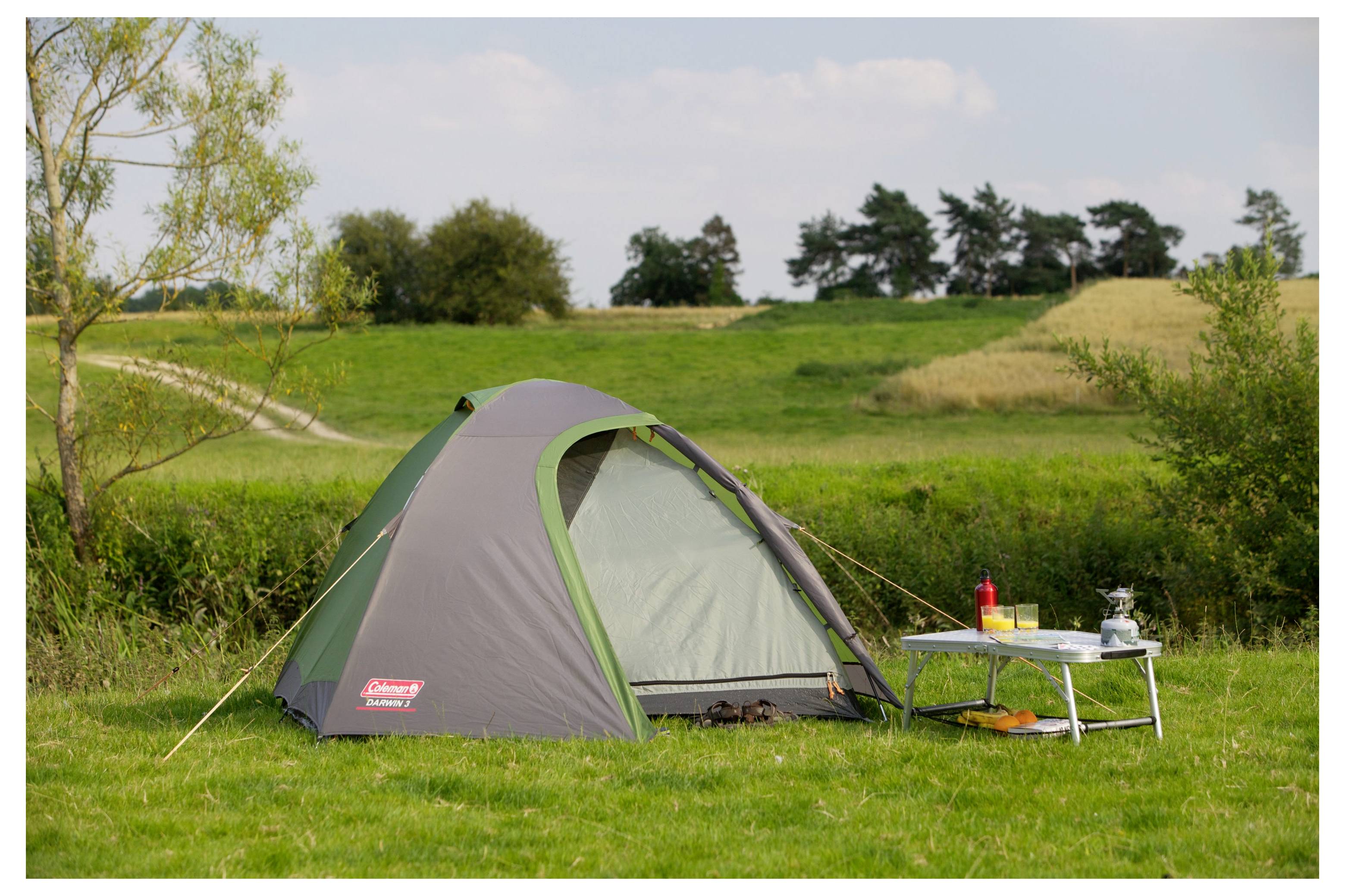 Ein kleines Campingzelt auf einer Wiese neben einem Klapptisch mit Kochutensilien, in einer ruhigen ländlichen Landschaft.