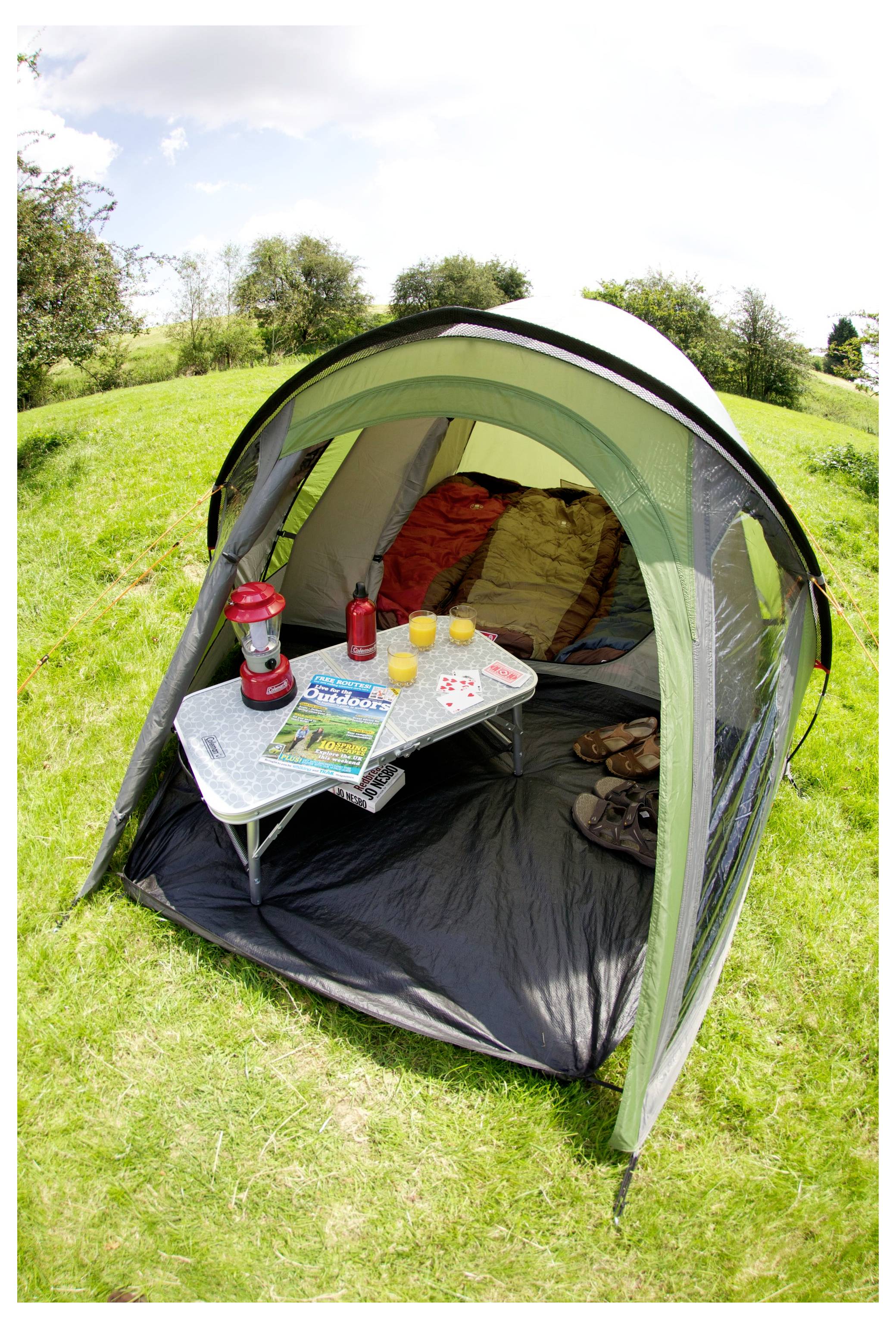Kleines Campingzelt auf einer Wiese mit Schlafsäcken im Inneren und einem Tisch außen, auf dem eine Laterne, Getränke und eine Karte oder ein Reiseführer stehen.