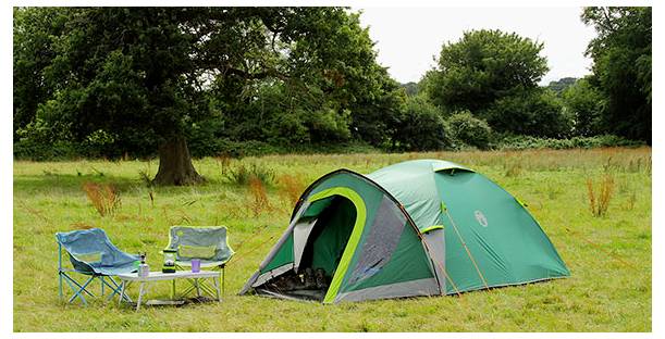Grünes Campingzelt auf einer Wiese aufgebaut mit zwei Klappstühlen und einem kleinen Tisch, umgeben von Bäumen.