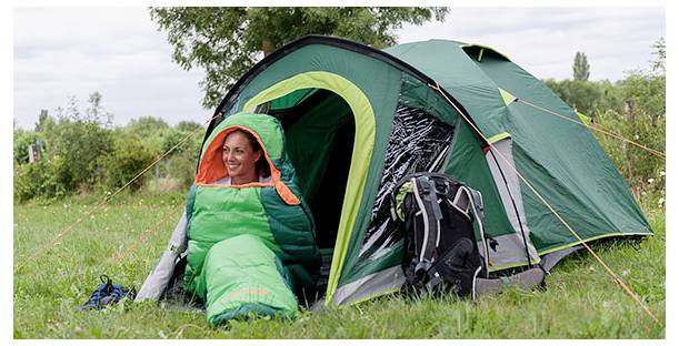 Eine Frau, die aus einem grünen Schlafsack am Eingang eines aufgebauten grünen Campingzelts in einem Grünland lächelt, mit einem Rucksack in der Nähe.