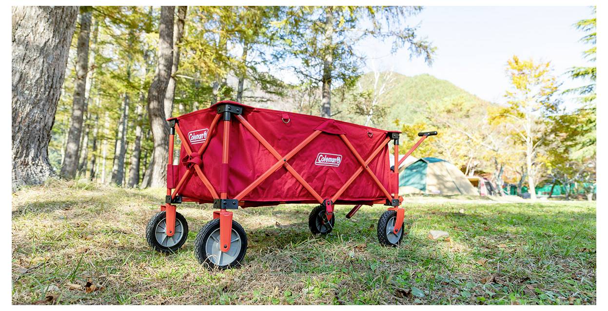 Roter Coleman-Klappwagen mit schwarzen Rädern, geparkt auf Gras an einem bewaldeten Campingplatz mit Zelten im Hintergrund.