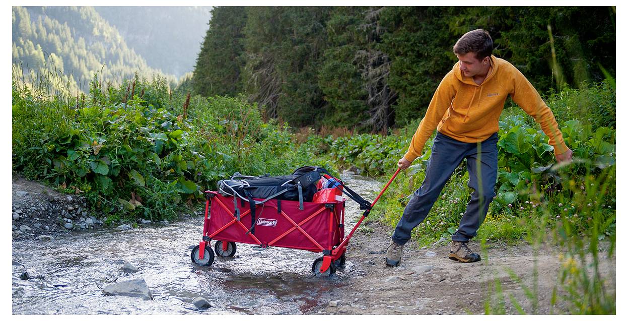 Ein Mann zieht einen roten Faltkarren, der mit Ausrüstung beladen ist, über einen felsigen Bach auf einem Waldweg in den Bergen.