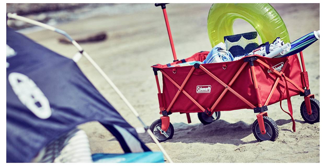 Roter Wagen auf einem sandigen Strand mit einem hellgrünen Schwimmring, Handtüchern und Flip-Flops, mit einem Segelboard oder Drachen im Vordergrund sichtbar.