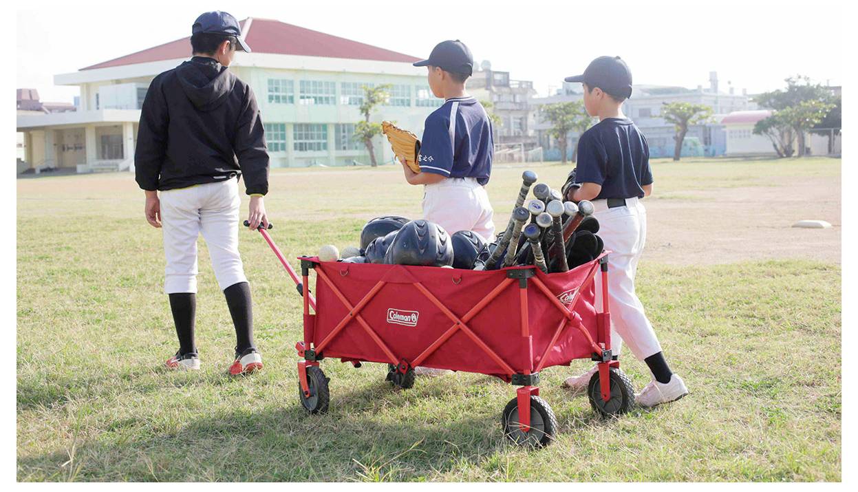 Drei junge Baseballspieler gehen über ein Feld und ziehen einen roten Wagen voller Schläger und Helme auf ein Schulgebäude zu.