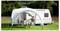 Eine Familie sitzt an einem Tisch unter einem Baldachin neben einem weißen Wohnwagen auf einem Campingplatz, mit Bäumen und Gras im Hintergrund.