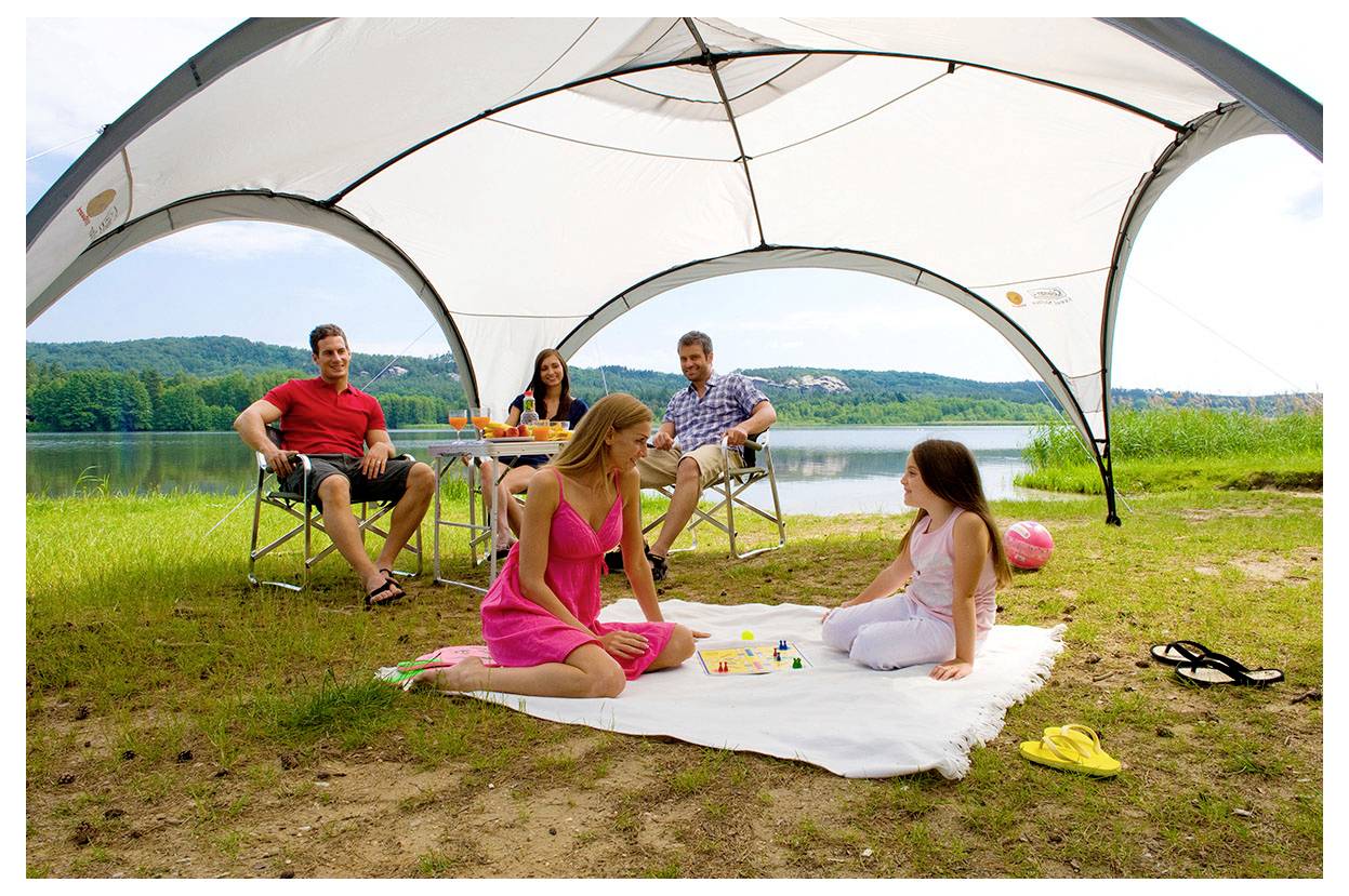 Familiencamp an einem See unter einem Zeltdach, mit entspannenden Erwachsenen in Stühlen und zwei Mädchen, die sich auf einer Decke im Vordergrund vergnügen.