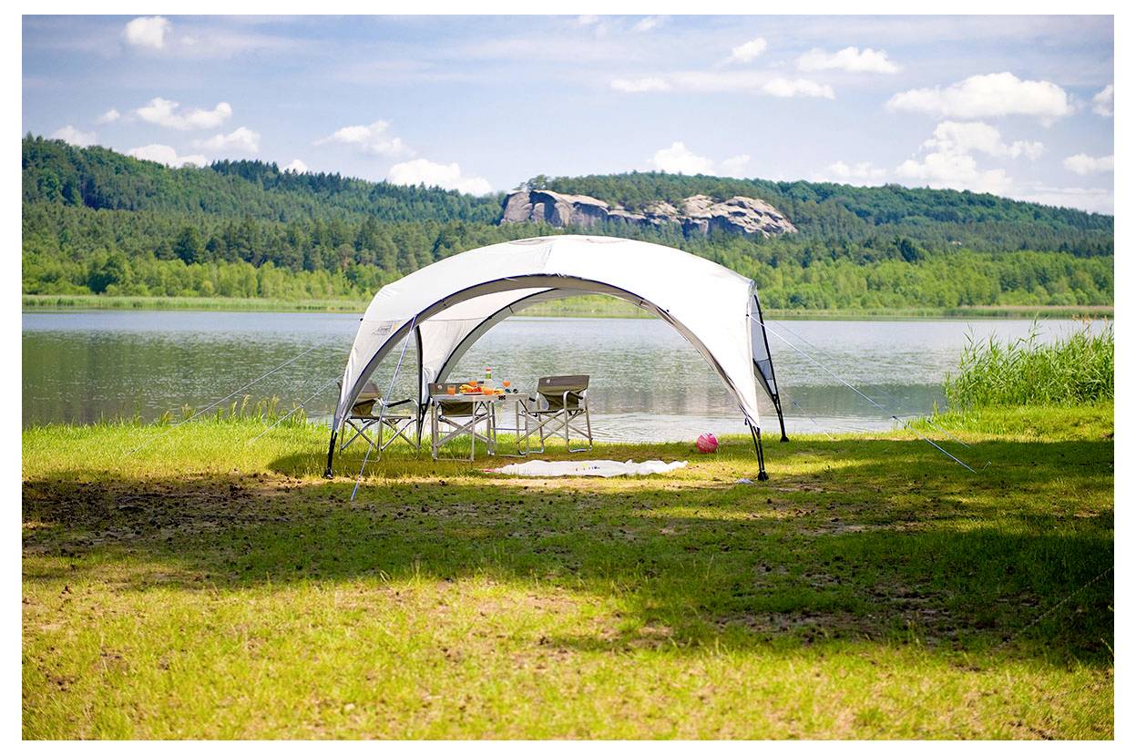 Weißes Pagodenzelt auf einem grasbewachsenen Seeufer aufgebaut, mit Klappstuhl und einem Tisch, die auf ruhiges Wasser und bewaldete Hügel im Hintergrund ausgerichtet sind.