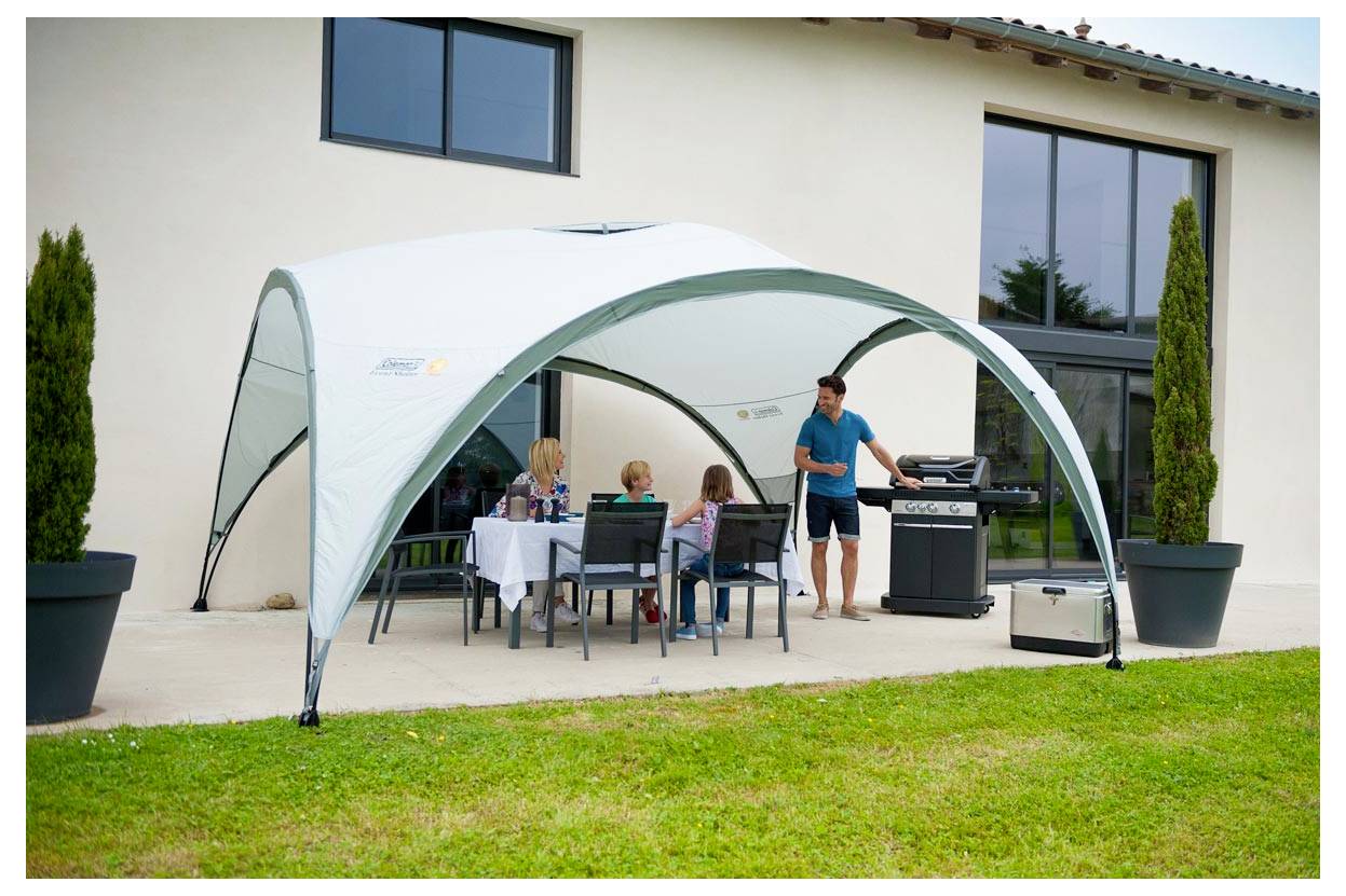 Menschen, die unter einem großen weißen Außenzelt neben einem modernen Haus sitzen, während ein Mann an einem Grill im Terrassenbereich steht.