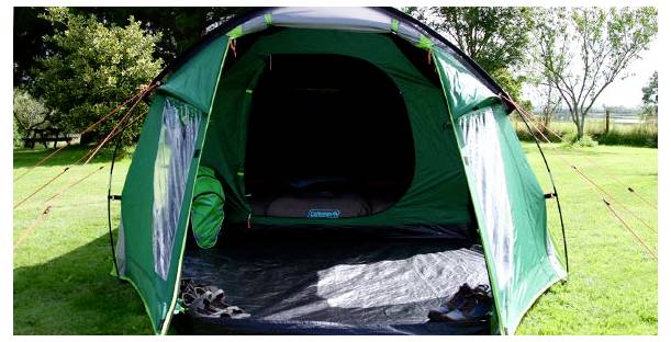 Offenes grünes Campingzelt mit Schuhen am Eingang, aufgestellt auf Gras in einem sonnigen Außengelände.
