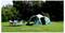 Campingaufbau auf einem grasbewachsenen Rasen mit einem grünen Kuppelzelt, Klappstühlen und einem kleinen Tisch unter Bäumen.