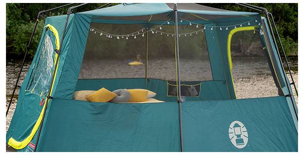 Türkisfarbenes Campingzelt mit Bildschirmfront, beleuchteten Lichterketten und Kissen im Inneren, das im Freien in der Nähe von Wasser aufgestellt ist.