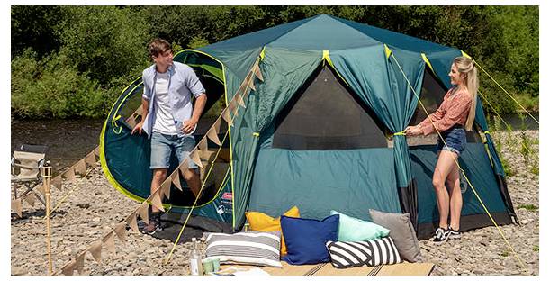 Ein Mann und eine Frau bauen ein blaugrünes Campingzelt auf einem felsigen Campingplatz am Fluss auf, mit Picknickdecken und Kissen davor.