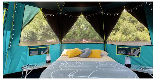 Innenraum eines glamping-Zelts mit einem Bett, Kissen, Nachttischen und Laternen mit Blick auf einen Fluss oder See und Bäume draußen.