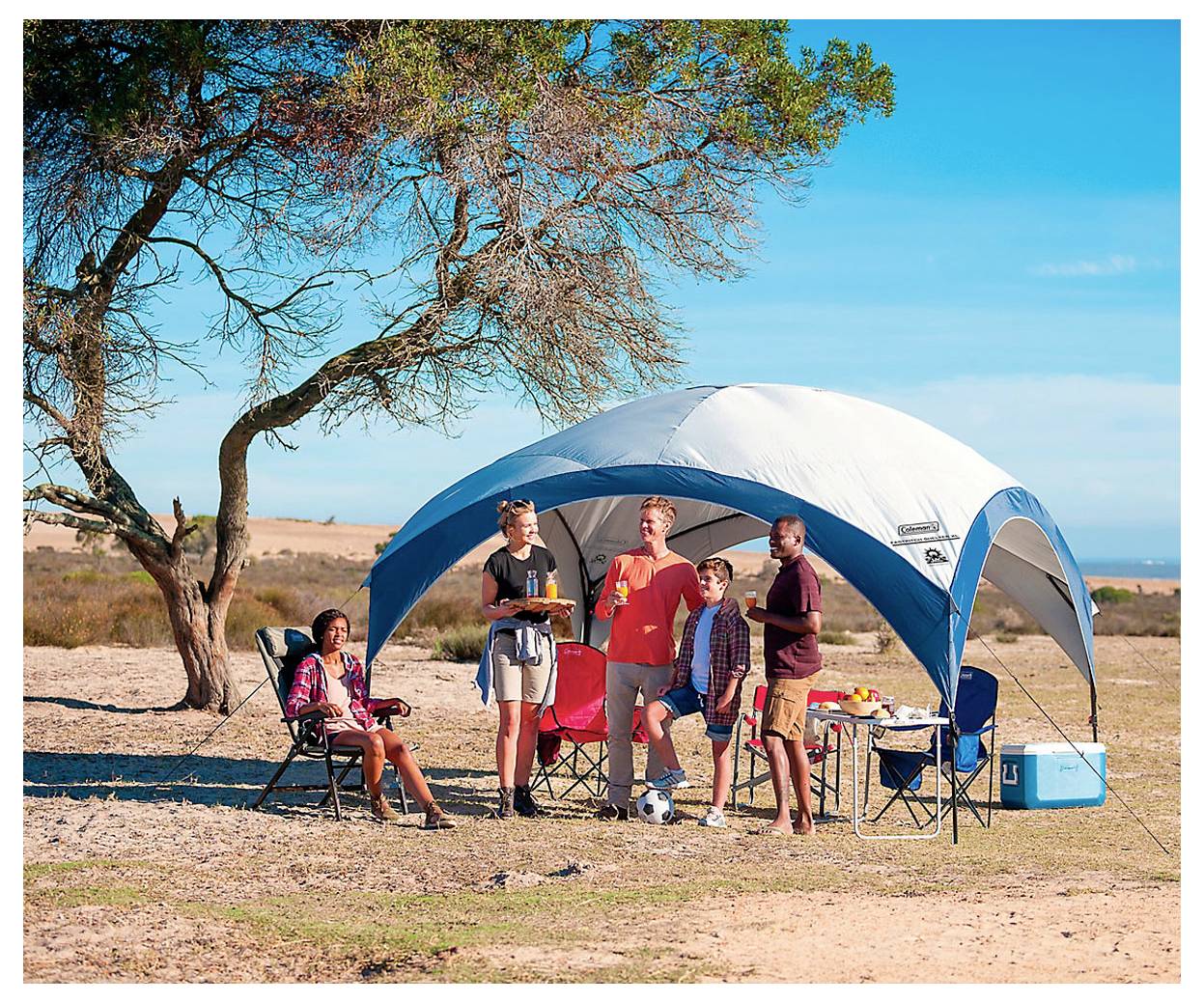 Vier Personen an einem Campingplatz unter einem großen Baldachzelt in der Nähe eines Baumes, mit Klappstühlen, Getränken und einer Kühlbox auf einer sonnigen, offenen Wiese.