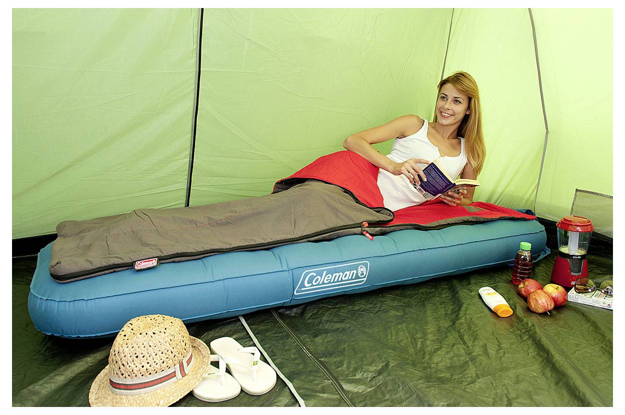 Frau, die sich in einem Schlafsack auf einer Luftmatratze in einem Zelt entspannt, hält ein Buch in den Händen, mit Campingausrüstung und Snacks in der Nähe.