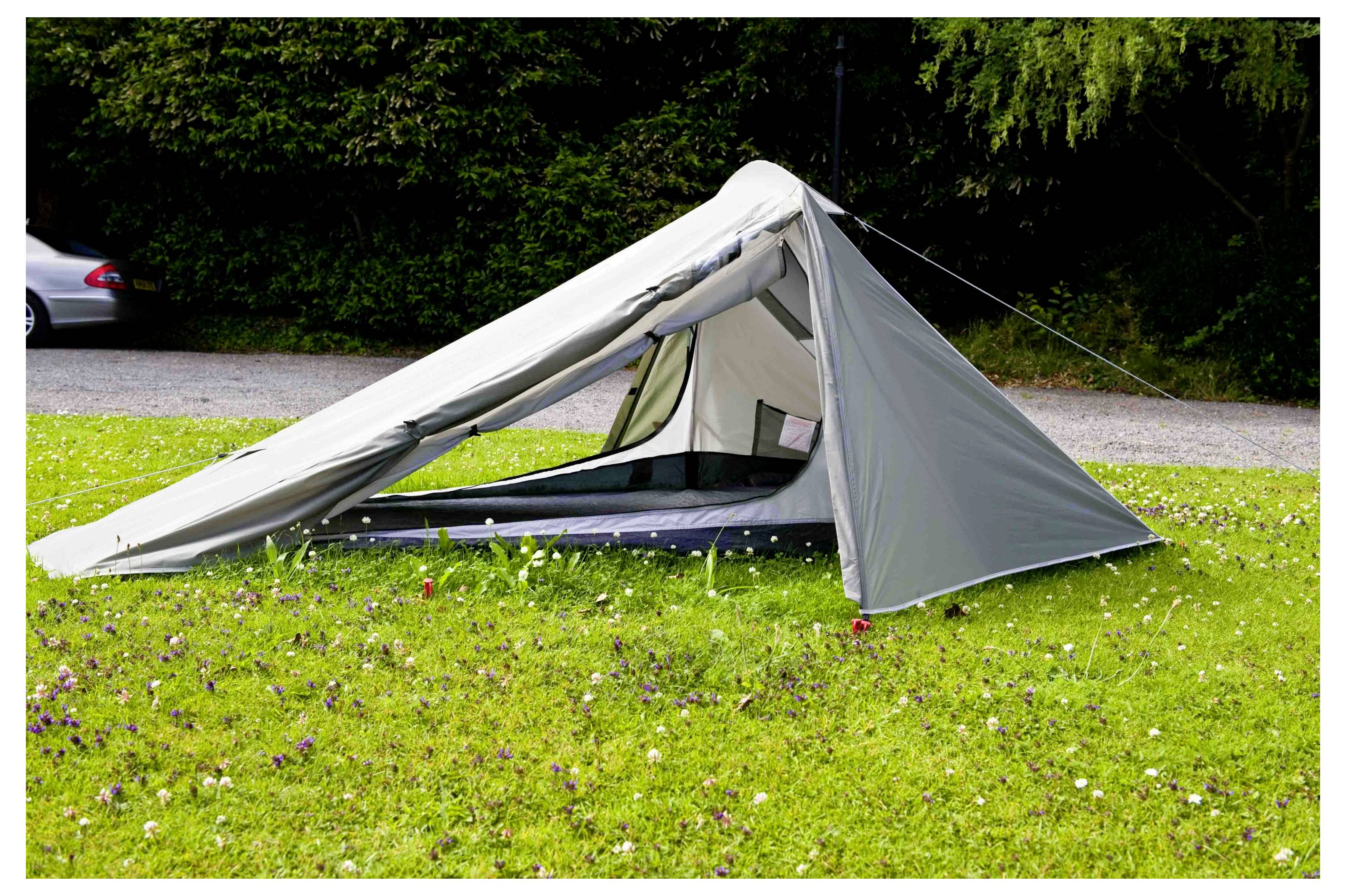 Ein graues Campingzelt auf einer Wiese aufgestellt mit Bäumen und einem geparkten Auto im Hintergrund.