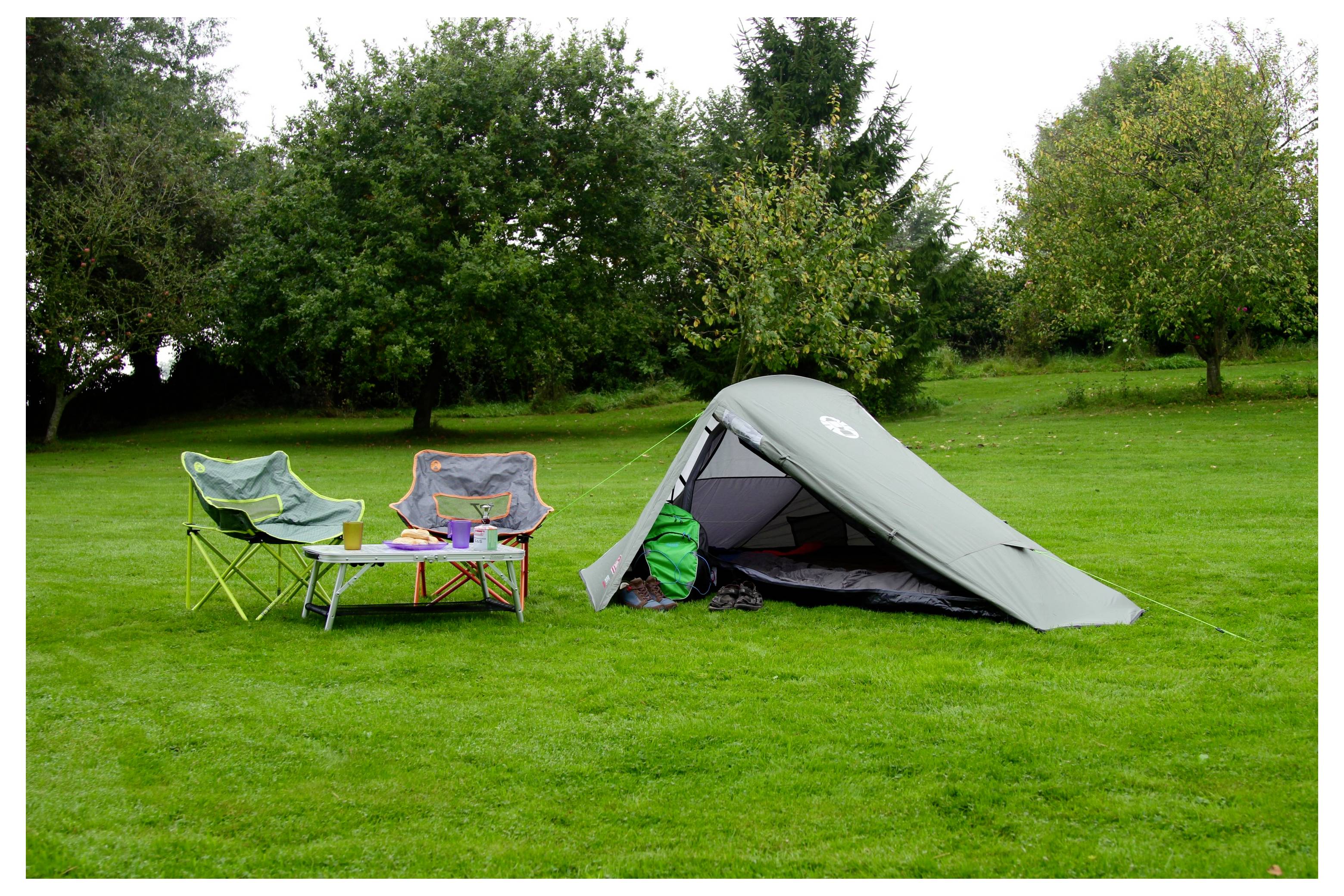 Ein kleines Campingset auf einem Grasplätzchen mit zwei Klappstühlen, einem niedrigen Tisch und einem grünen Zelt, das in der Nähe von Bäumen aufgestellt ist.