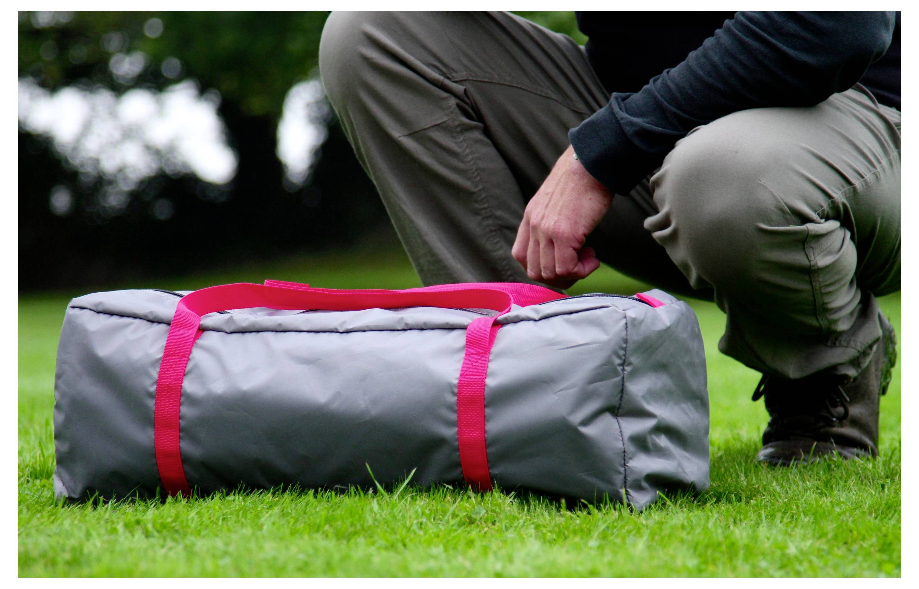 Person, die neben einer grauen Reisetasche mit roten Riemen auf Gras kniet, im Freien, bereit, sie zu öffnen oder zu packen.
