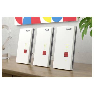 Drei weiße FRITZ! Wireless-Geräte stehen aufrecht auf einem Holzschreibtisch, jedes mit einer roten Taste und Signalleuchten, neben einer Pflanze.