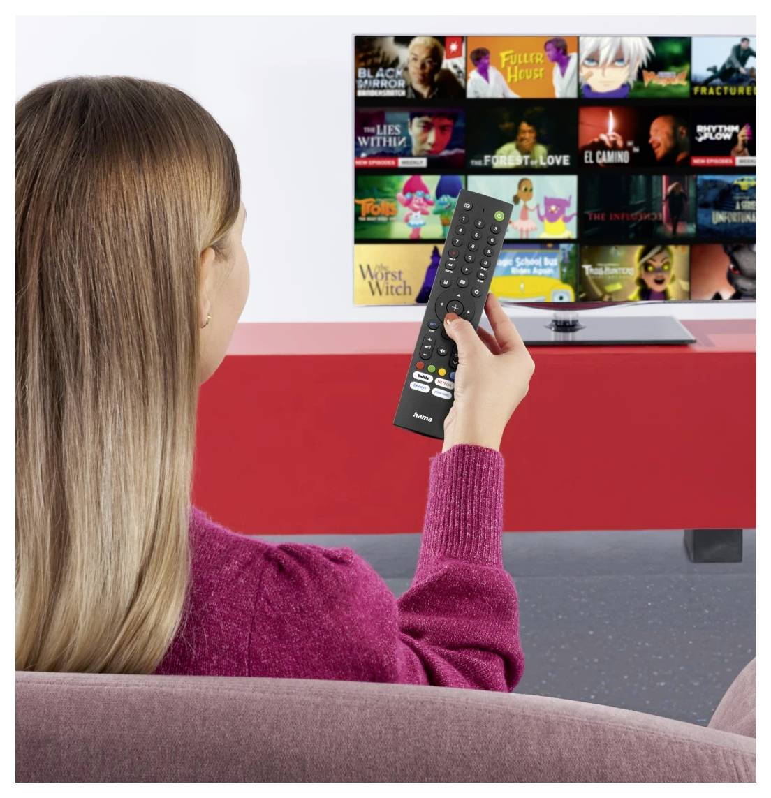 Frau auf einer Couch zeigt mit einer Fernbedienung auf einen Fernseher, der ein Streaming-App-Gitter mit Film- und Serien-Miniaturansichten anzeigt.