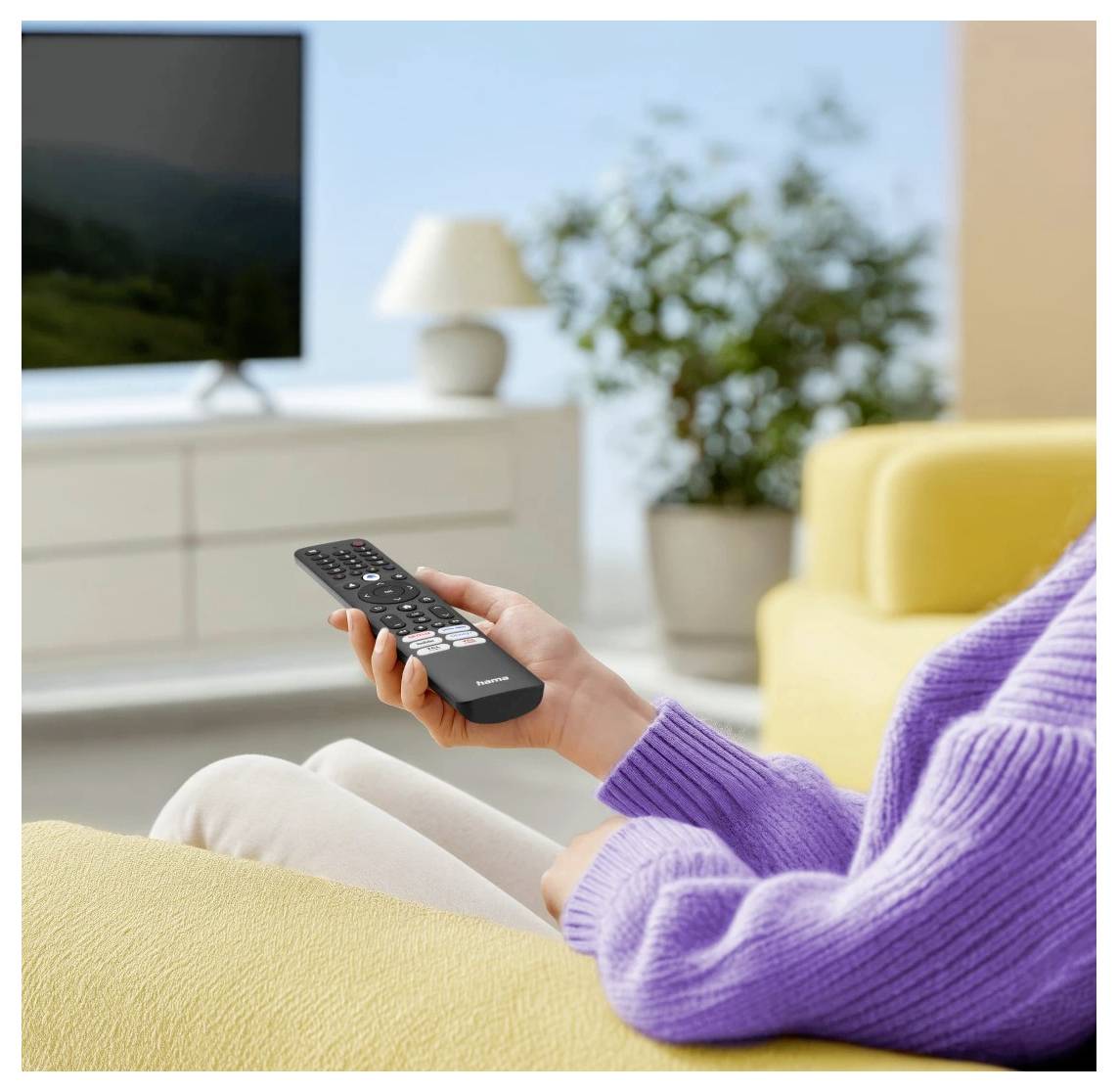 Eine Person in einem lila Pullover sitzt auf einem gelben Sofa und hält eine Fernbedienung in der Hand, während sie einen Fernseher in einem hellen Wohnzimmer anschaut.